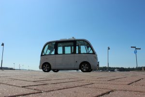 in the front stone pavement, in the middle an automated electric shuttle, in the background clear blue sky
